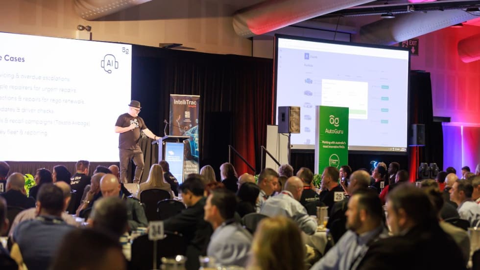 A presenter speaks on stage at a conference, addressing an audience seated at round tables, with large screens displaying presentation slides in the background.