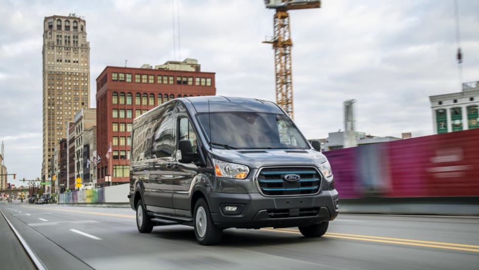 A gray 2022 Ford E-Transit driving on a city road with construction in the background.