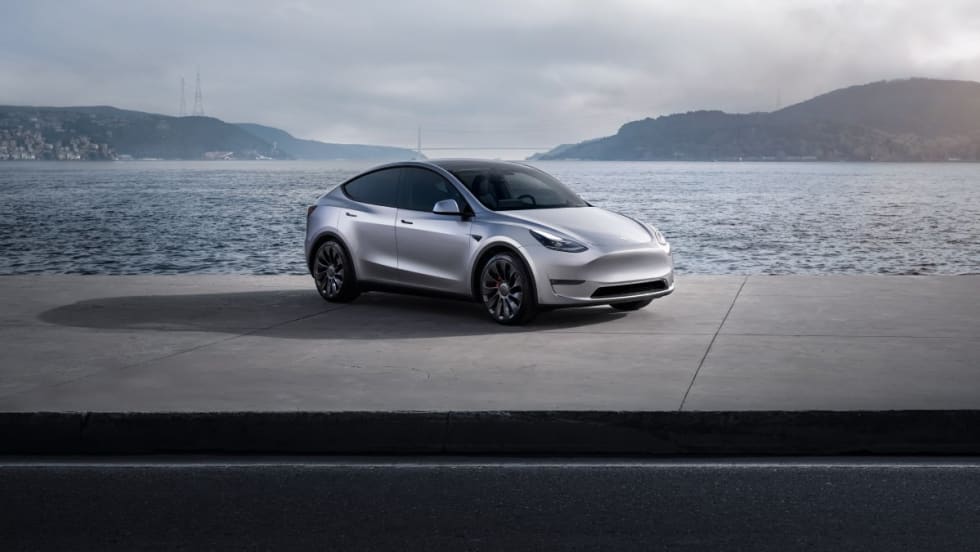 A silver Tesla Model Y parked in front of a body of water and hills in the background.