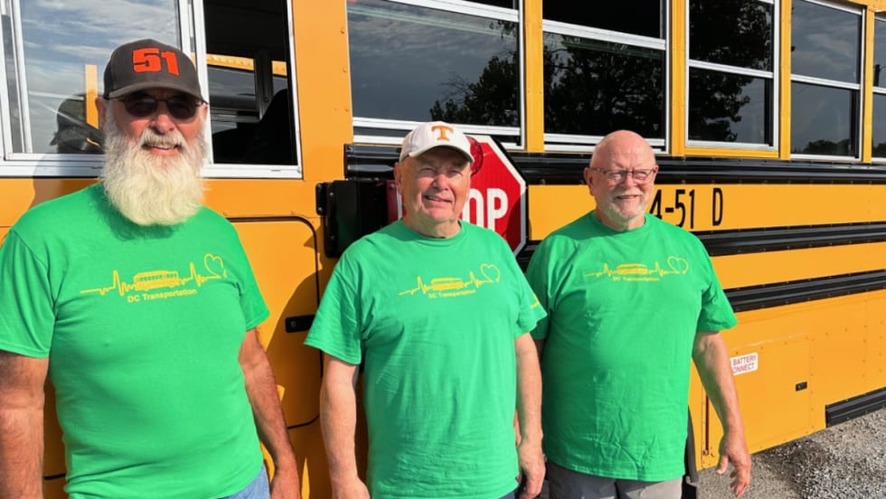Three men in green shirts pose next to a school bus.
