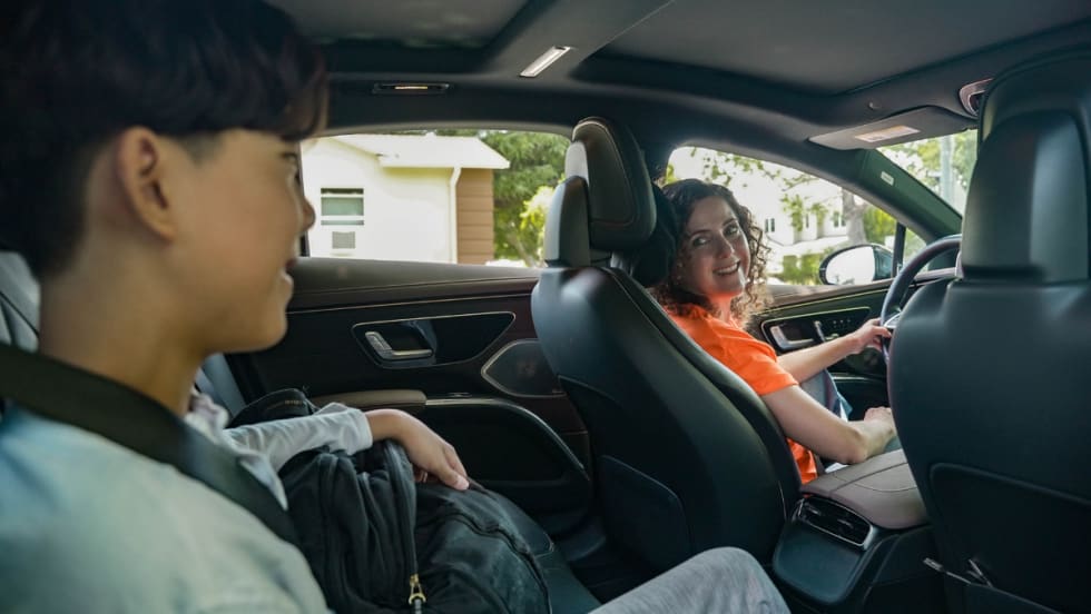 Female HopSkipDrive CareDriver in an orange shirt driving a student to school in a car.