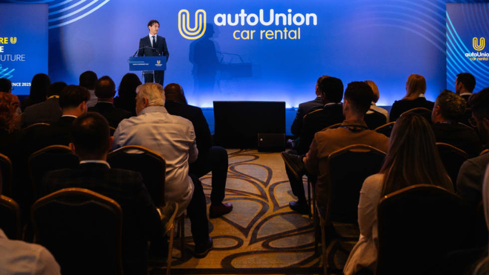 AutoUnion COO speaks to audience in front of wide blue screen with company logo on stage.
