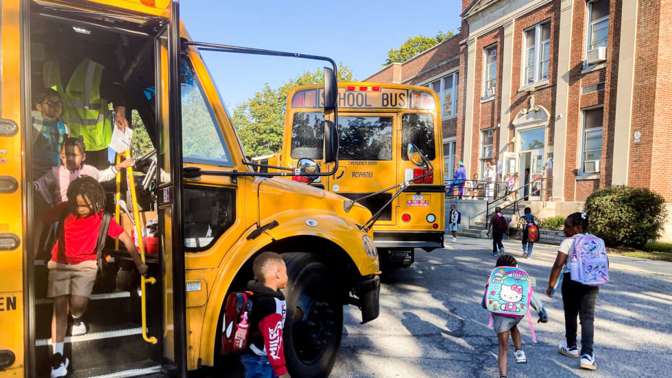 Students with backpacks exit school buses and walk toward a school building as transportation staff assist with afternoon drop-off.