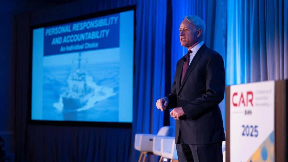 Commander Lippold on stage in front of projected photo of USS Cole.