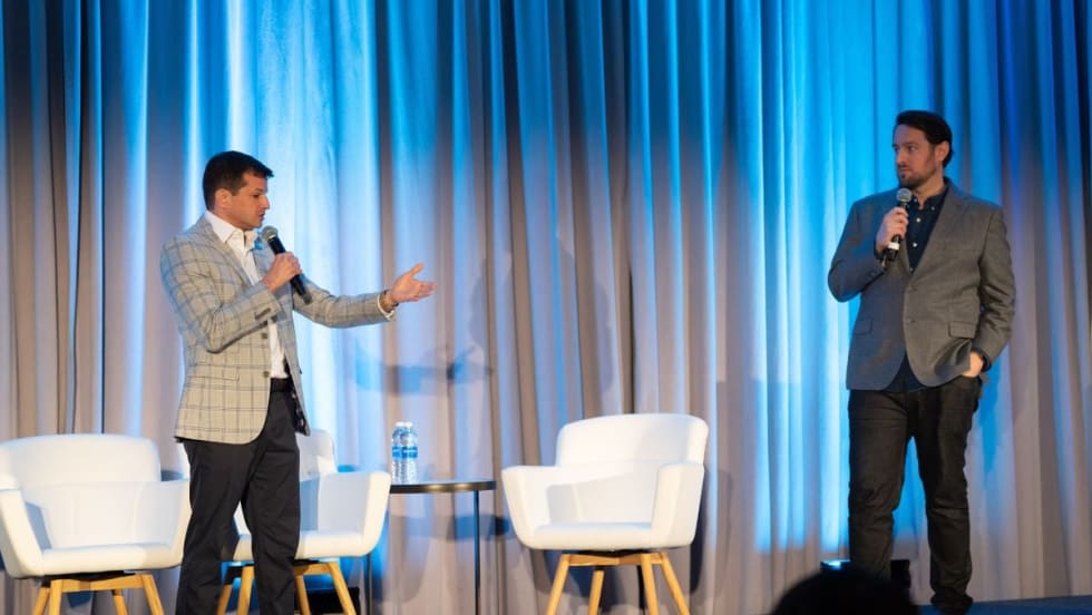 Two speakers standing on a stage talking at each other with a white chair between them.