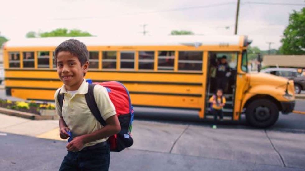 a child walks on the sidewalk with a school bus behind him