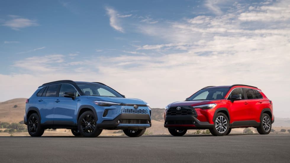 The 2026 Toyota Corolla Cross and its hybrid counterpart sit next to each other on the road with a blue sky background behind them. 