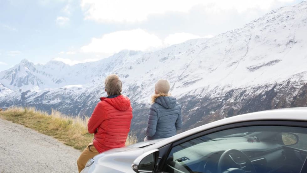 A couple sitting on the hood of a rental car looks at the scenic snowy mountain range.