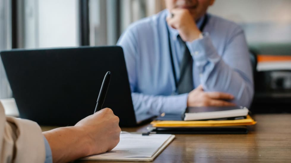 Two people facing each other at a desk, the person in the foreground is filling out papers.