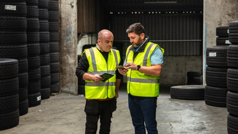 Two workers in safety vests look at a tablet and phone in a tire warehouse.