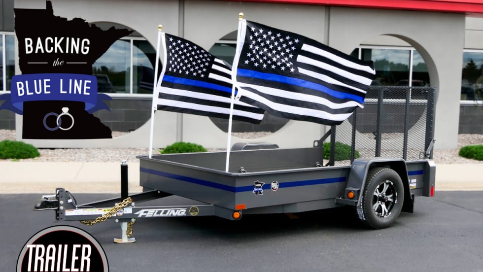 A dark gray dropdeck utility trailer with a thin blue line is shown, with 2 Thin Blue Line flags hanging off the back.