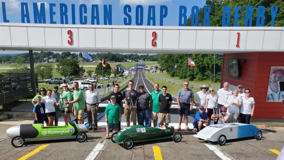 A group of participants and organizers stand behind three colorful soap box cars at the starting line of the All-American Soap Box Derby track in Akron, Ohio, celebrating the launch.