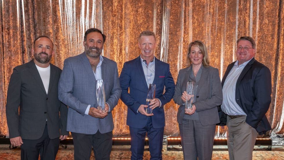 Five people stand against a gold background holding awards