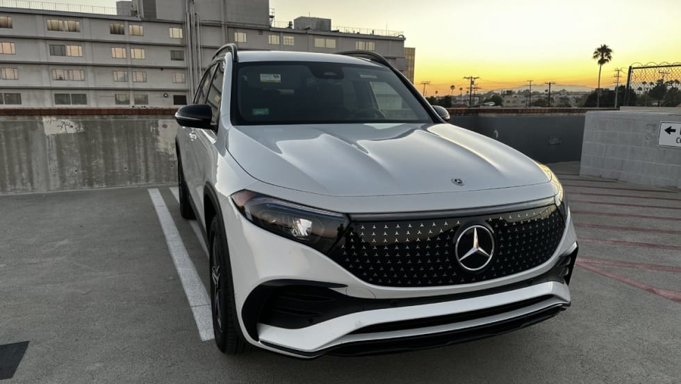 The front of a white Mercedes-Benz EQB 250+ in a parking space with a sunset in the background.