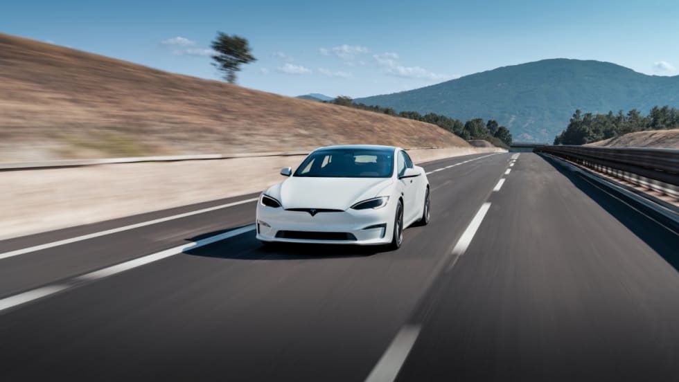 A white Tesla Model S driving down a paved road with hills and mountain in background on a sunny day.
