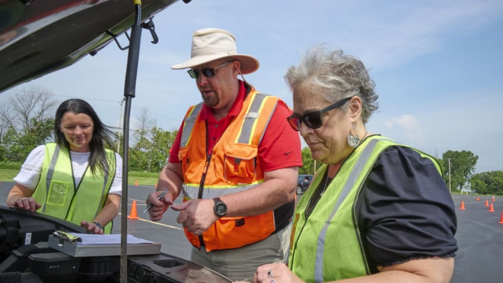 A man in an orange vest shows two women in green vests items under the hood of a vehicle.