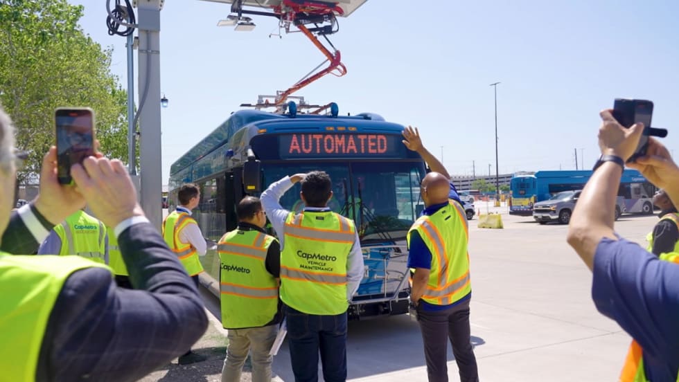 Image of a blue CapMetro bus at an overhead charging station as employees watch.