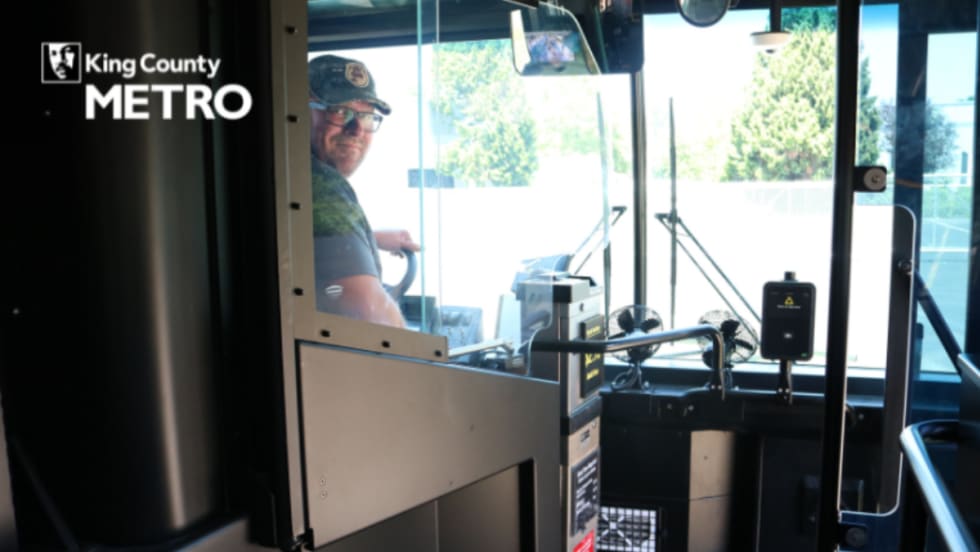 Image of a bus operator posing from the front seat of a public bus, behind a clear partition.