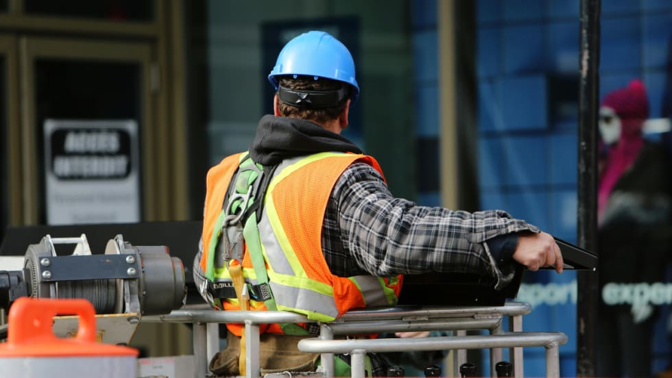 The back of a construction worker wearing a blue hard hat and an orange visibility vest.