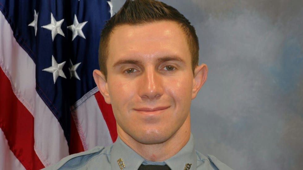 Police officer in light blue uniform standing in front of American flag