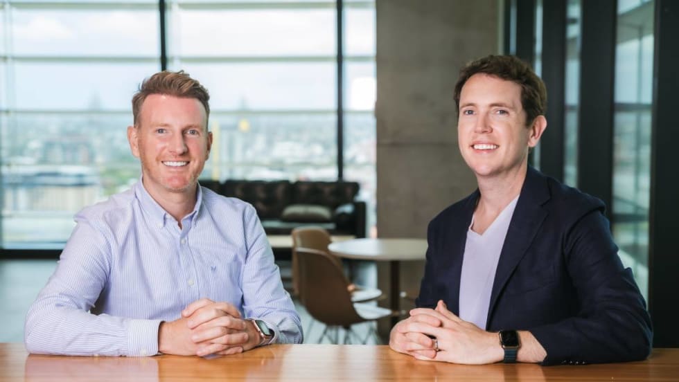 Two corporate-looking young men smile along a gleaming wooden conference table.