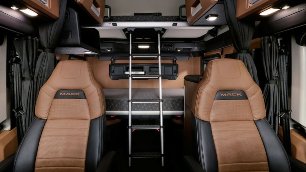 Interior view of the Mack Pioneer sleeper cab, showing two leather seats, an upper bunk with a ladder, overhead storage compartments, and integrated lighting.