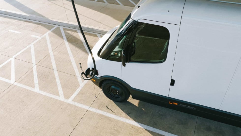 A white Rivian commercial van is charging in a parking lot. 