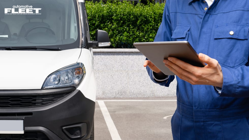 A man in a blue mechanic jumper holds a tablet at chest level in front of a white van.