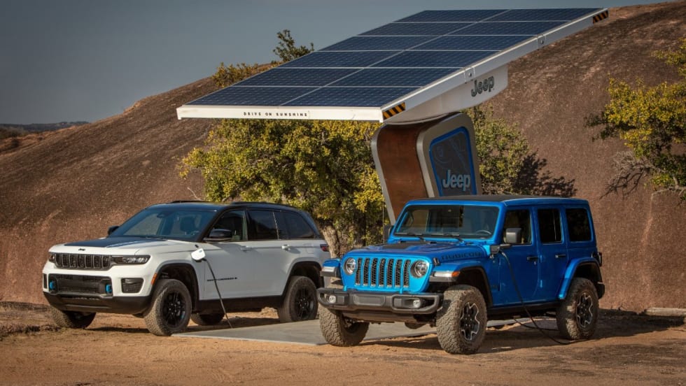 A white 2022 Jeep Grand Cherokee and a cobalt blue 2022 Jeep Wrangler recharge at a Jeep charge station in a desert.