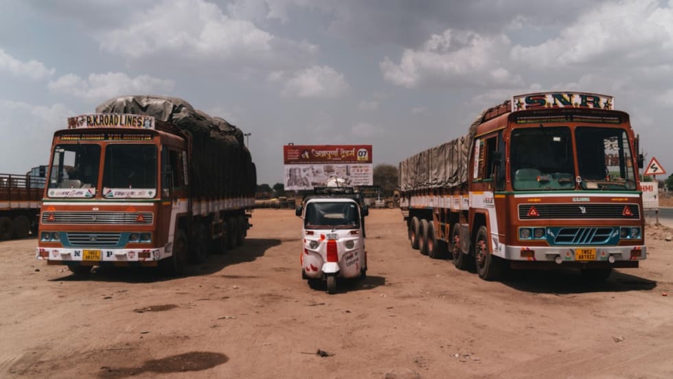 A rickshaw parked between two red transit buses in India.