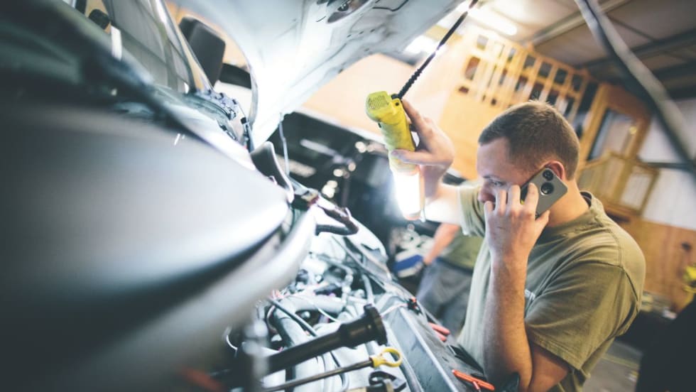 A man holding a light and a phone looking at the engine of a vehicle.