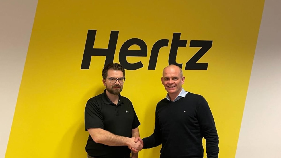 Two executives shaking hands in front of bold yellow Hertz sign.