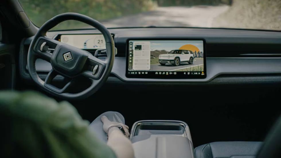 Interior view of a Rivian vehicle showing steering wheel and digital display screen with vehicle interface, representing autonomous technology development.