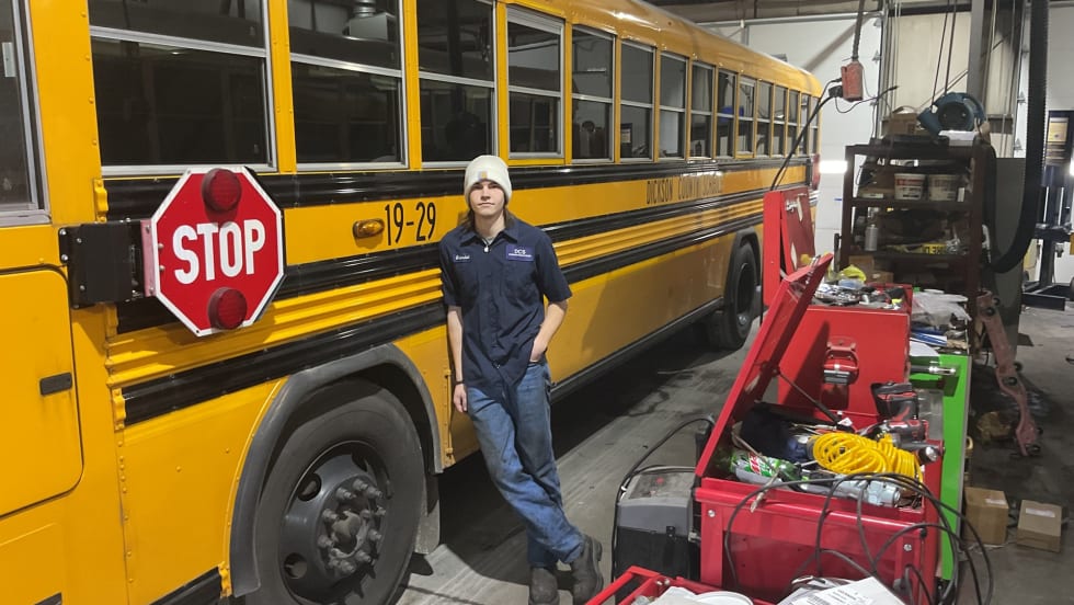 a young man leans against a school bus in a maintenance garage