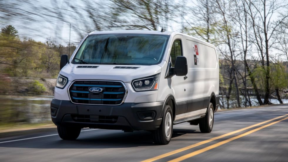 A Ford E-Transit van driving down a road with trees in the background.