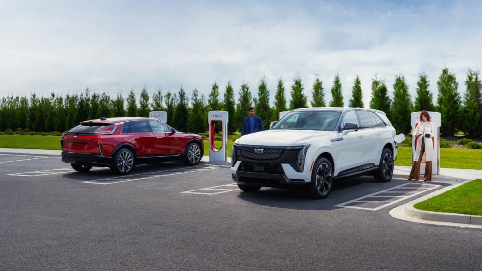 A red suv and a white suv parked in front of tesla superchargers with trees in the background.