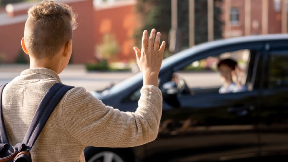 A young boy wearing a backpack waves at an adult sitting in a car.