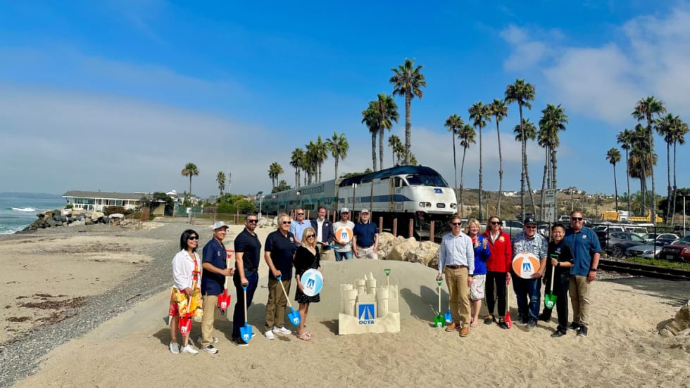 A group of people stand on a beach with a rail car in the background.