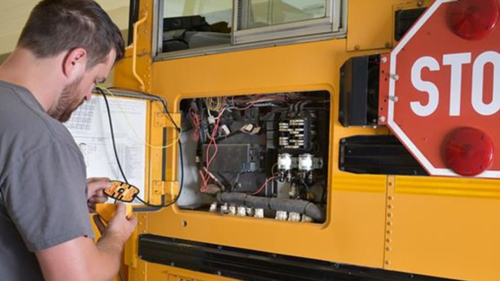 A man in a grey t-shirt works on the electrical components to a school bus stop arm.