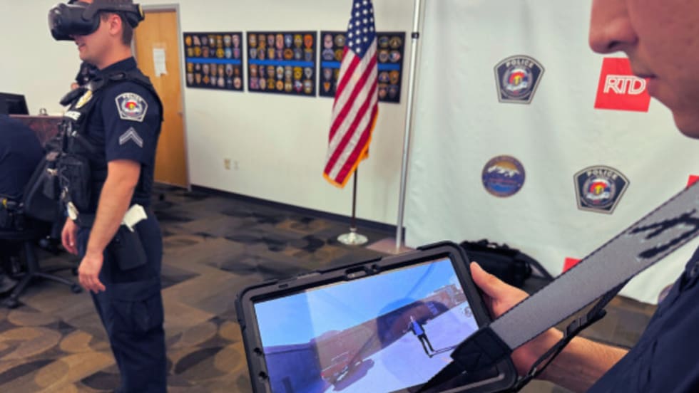 A RTD transit police offer holds up a tablet showing virtual reality training while an officer in the background stands with a VR headset on.