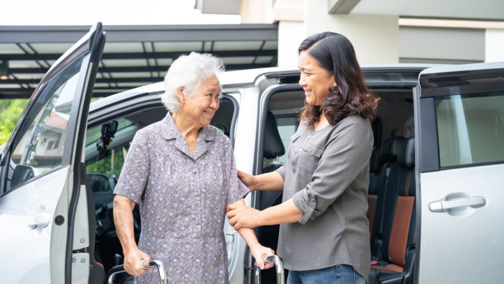 A woman helps an elderly woman with a walker out of van.