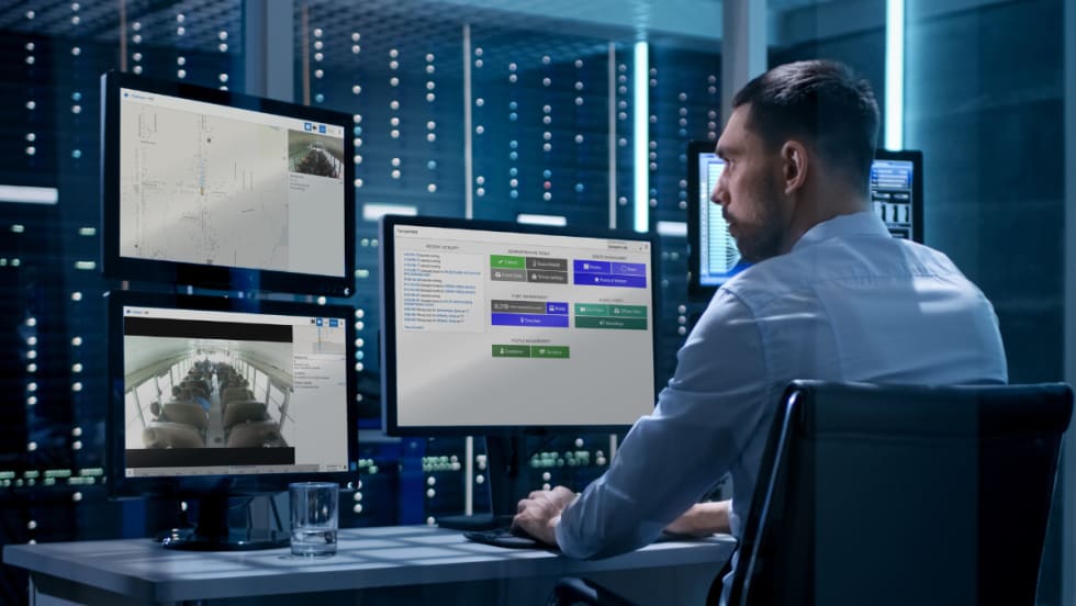A man sits at a desk with three computer monitors displaying Transportant's platform.