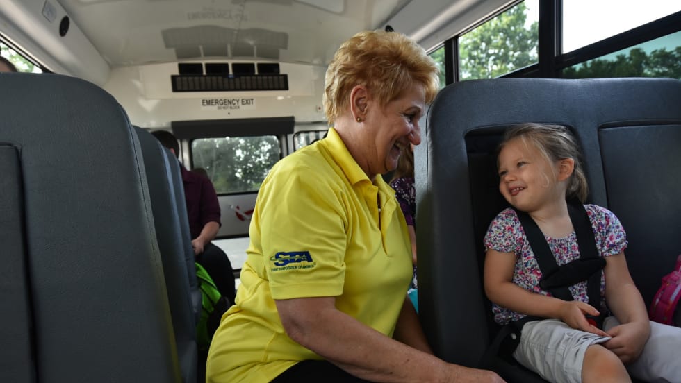 A school bus driver from Student Transportation of America smiles and talks with a young girl buckled safely into her seat on the bus.