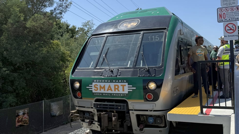 A SMART (Sonoma-Marin Area Rail Transit) train stopped at a platform during a station opening event, with people standing beside the train and signage posted near the edge of the platform.