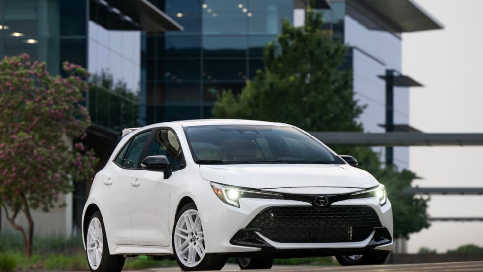 An Ice Cap Toyota Corolla Hatchback drives on the road with a glass building behind it. 