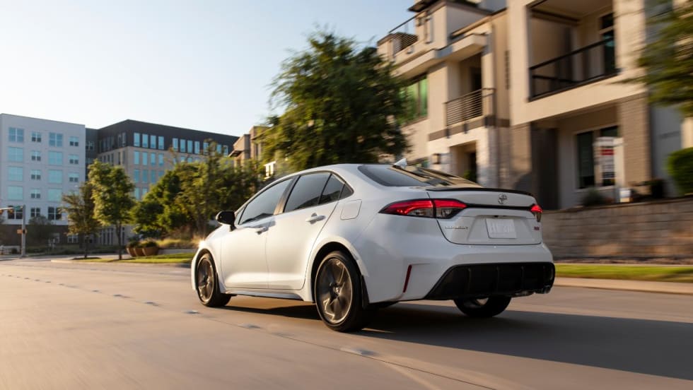 A 2026 Toyota Corolla drives on the road towards a glass building.