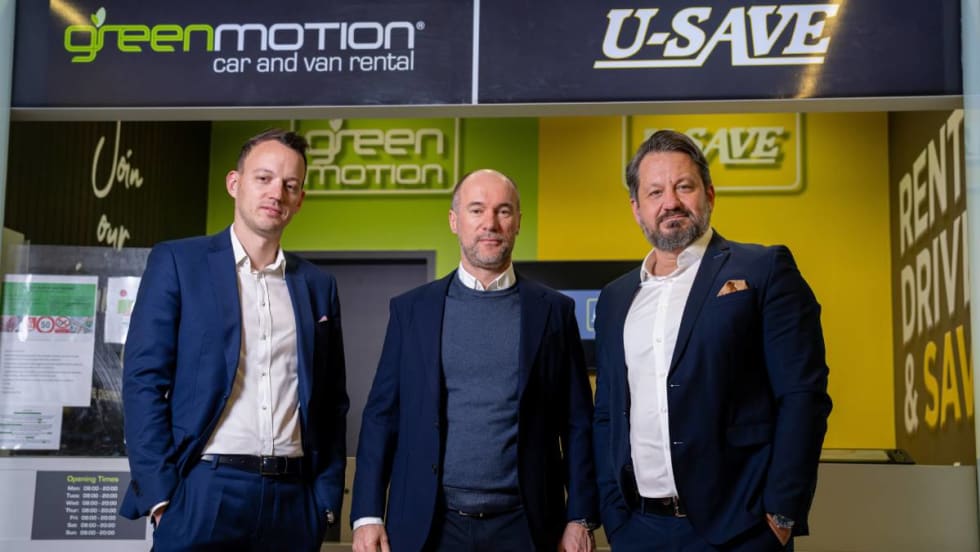 Three U-Save executives in suits pose in front of a rental car counter.