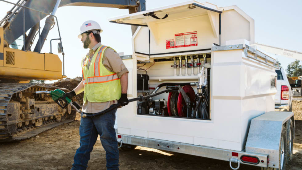 A worker in a yellow vest brings a fuel nozzle from a mobile fuel tank to a piece of equipment on a jobsite.