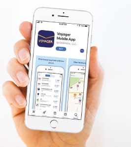 A hand holds a smartphone displaying the Voyager Mobile App page in the Apple App Store. The screen shows the app icon, blue “Get” button, and app interface previews featuring fuel price locations and station maps.