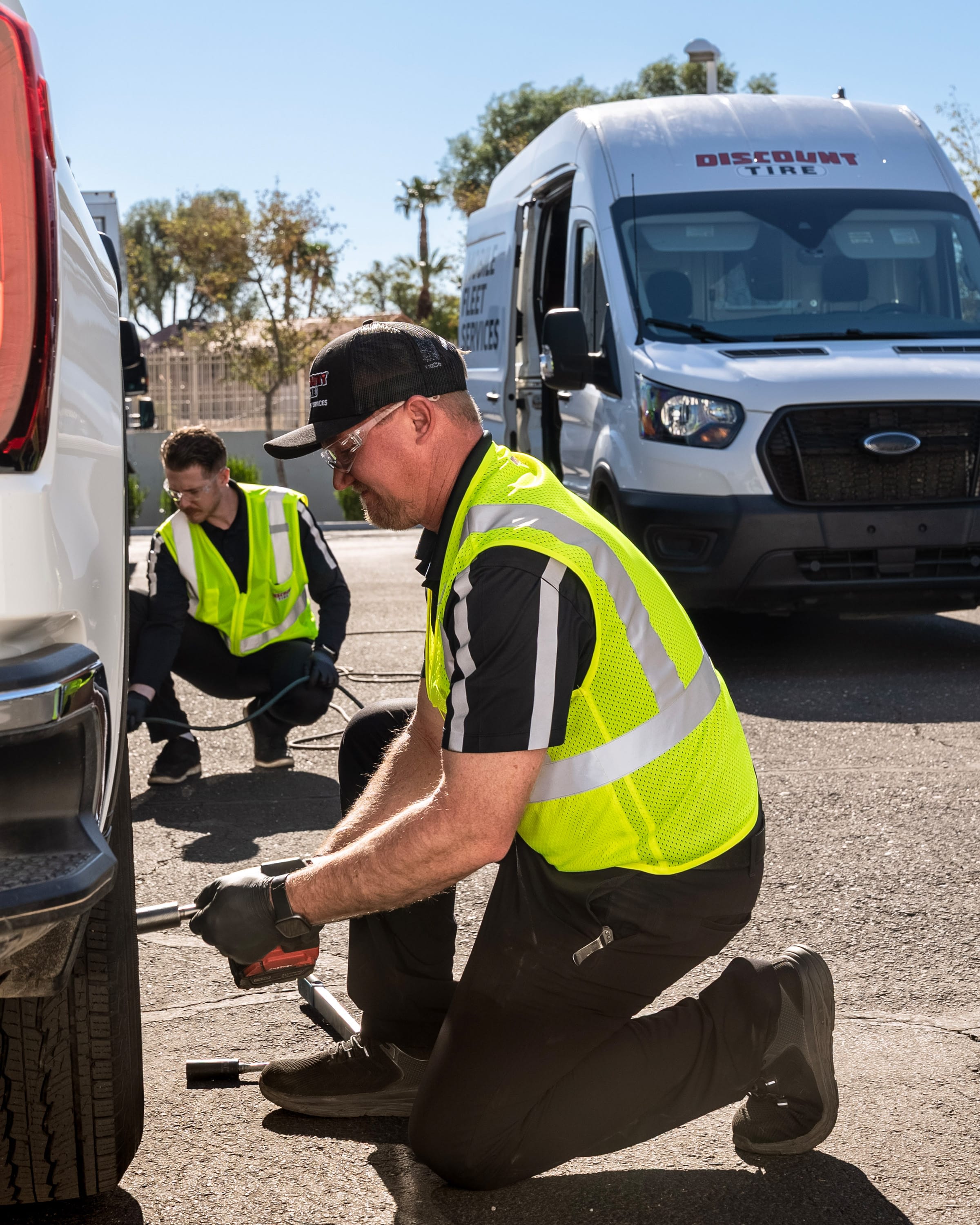 Discount Tire Fleet Services mobile services in high-visibility vests change a flat tire beside service vans on a sunny day, kneeling and using tools to secure the wheel.